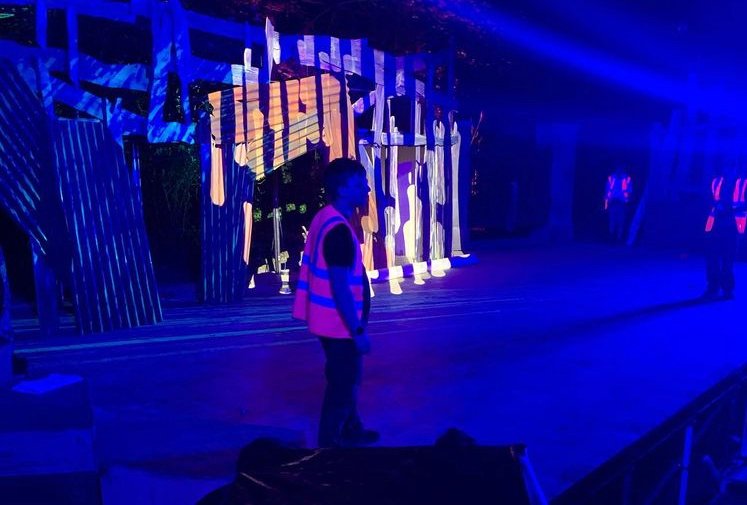 A young boy stands on a stage with colorful, illuminated wooden props and structures behind him, in a dimly lit setting with blue lighting.