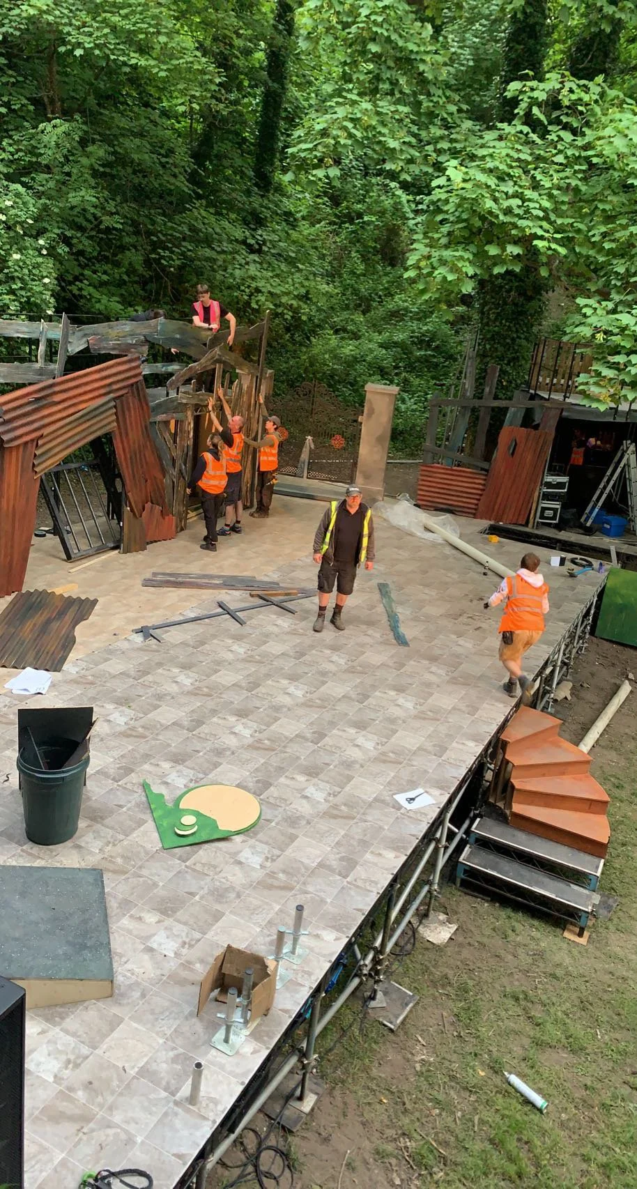 Construction workers onsite, some wearing orange safety vests, assembling or repairing a wooden outdoor structure surrounded by trees.