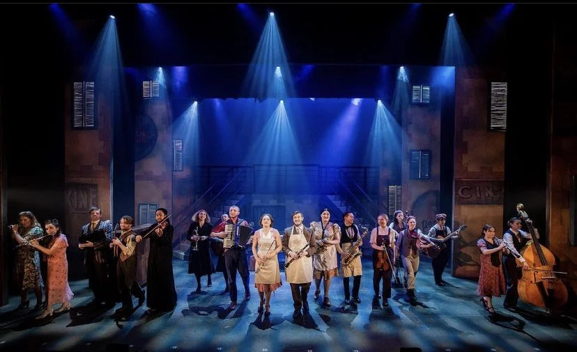 Group of performers on stage with musical instruments, stage lights, and set design in the background, during a theatrical performance.