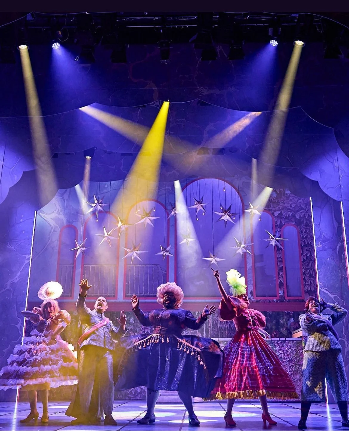 Theatrical stage with five performers in colorful costumes under bright yellow and purple spotlights, decorated with hanging star decorations, set against an elaborate backdrop.