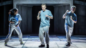 Three male dancers performing on stage with dark background and industrial set pieces.