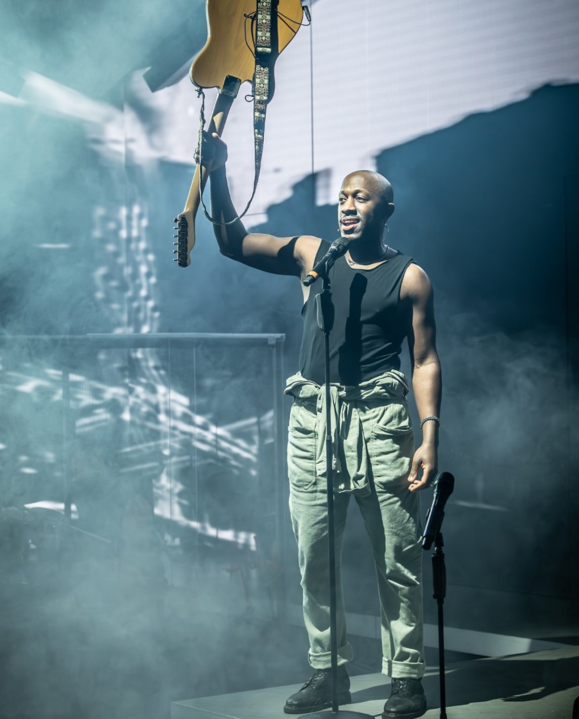 A person standing on stage holding an electric guitar above their head while speaking or singing into a microphone, with stage lights and fog in the background.