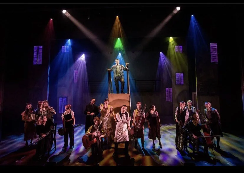 A theatrical stage scene with a performer in a striped blazer standing on an elevated platform, surrounded by a group of musicians and actors in period costumes. Colored stage lights illuminate the scene.