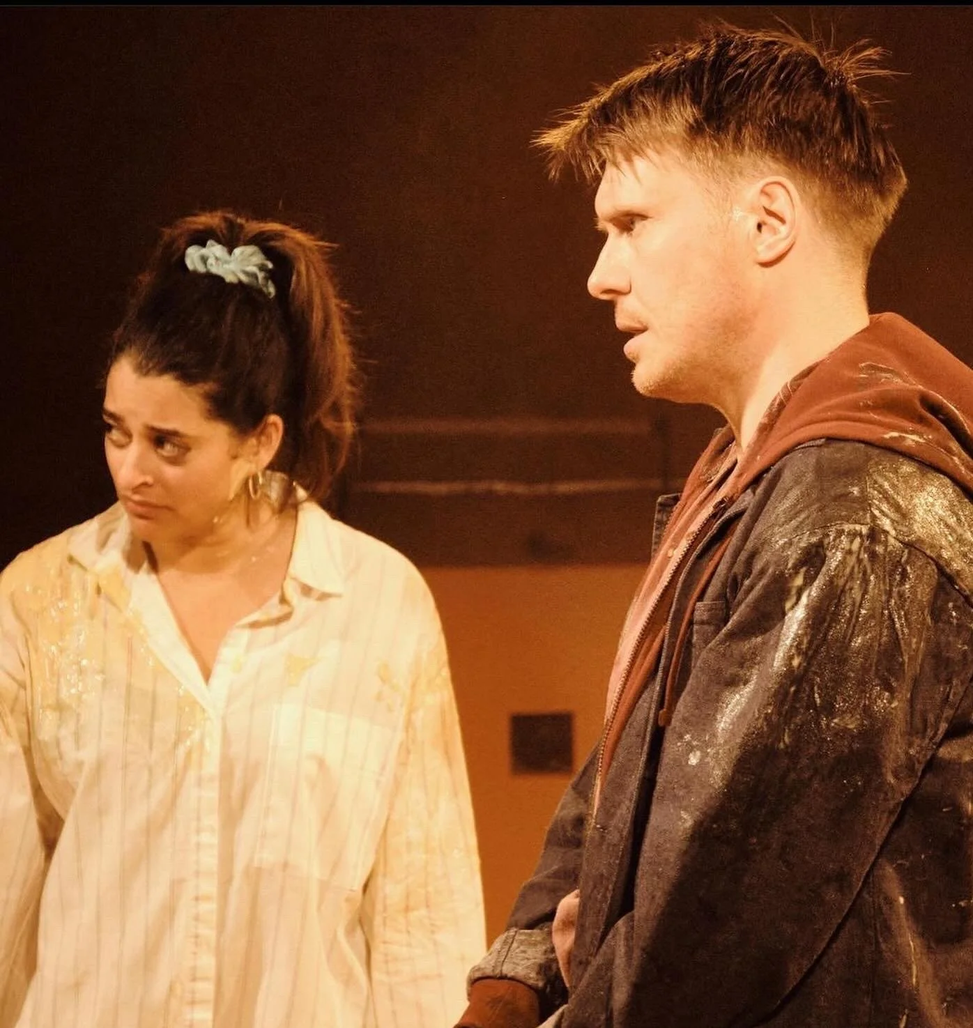 A woman with a high ponytail and a yellow-striped shirt looking sad or disappointed, standing next to a man with short hair, wearing a brown jacket and hoodie, both looking serious against a blurred warm-colored background.