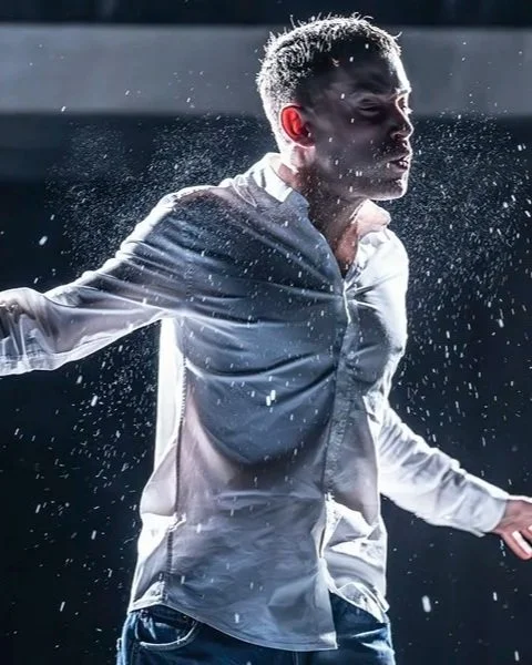 A young man wearing a white dress shirt is splashing water on himself, with water droplets visible around him, and a dark background.