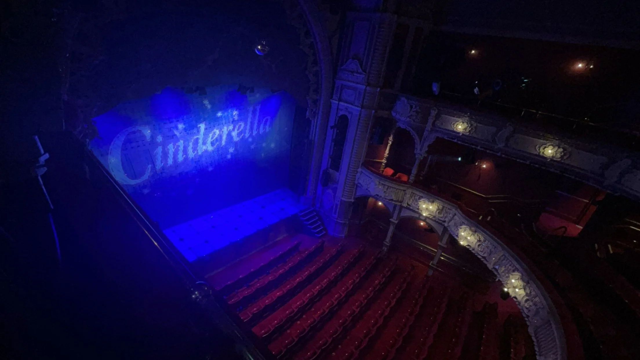 Empty theater stage with a large illuminated sign that reads 'Cinderella', red theater seats, ornate balconies, and a dark, dimly lit ambiance.