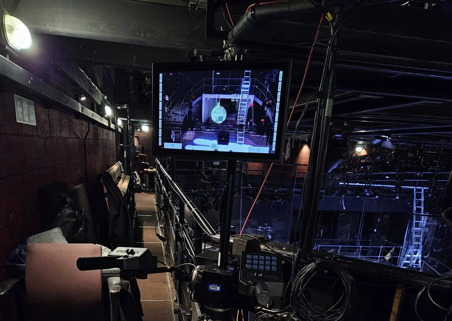 View of a theater stage with scaffolding and equipment, seen from the side of the seating area, with a monitor displaying a view of the stage setup.