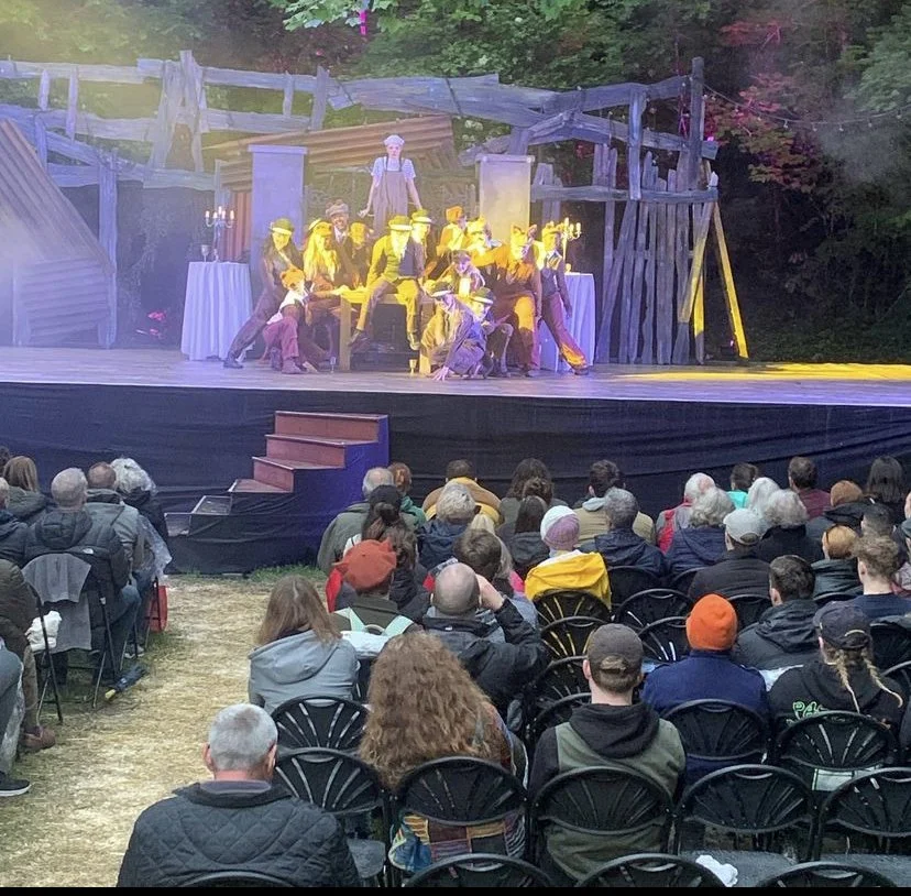 Actors performing on an outdoor stage at a theater in front of a seated audience, with a rustic, wooded backdrop.