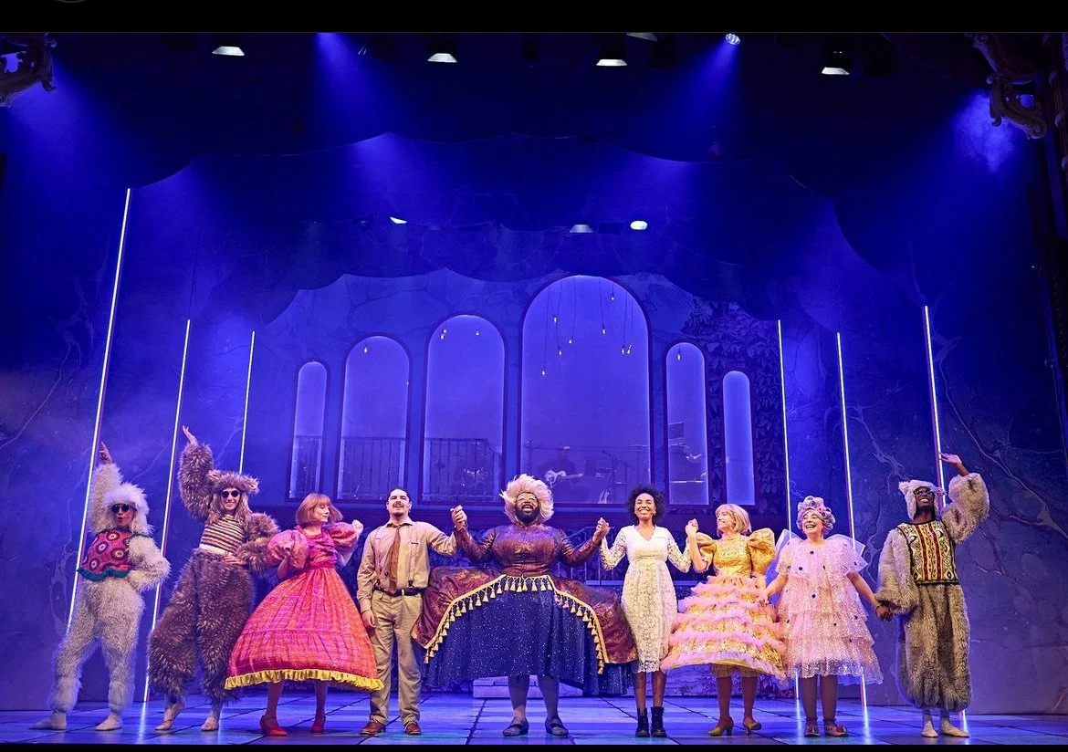 Stage performance featuring a diverse cast of actors in colorful costumes, holding hands and taking a bow on a lit stage with a dreamy, whimsical backdrop.