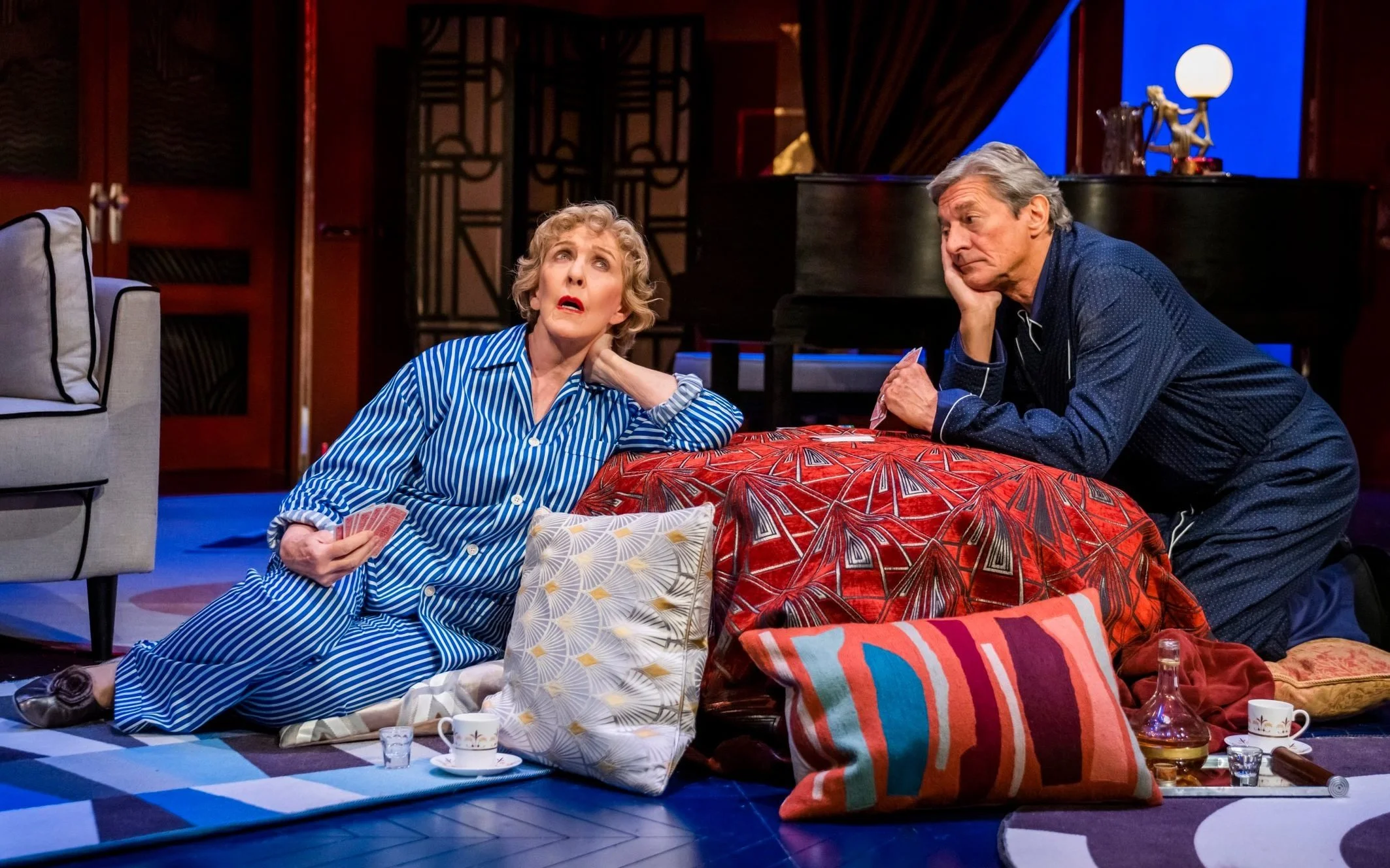 Older woman and man with gray hair, dressed in pajamas, sitting on a cozy floor in a living room, surrounded by pillows, with a teapot and cups, engaged in a dramatic conversation.