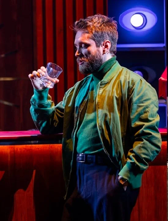 Man with a beard in a green jacket and green shirt holding an empty glass in a bar or club setting.