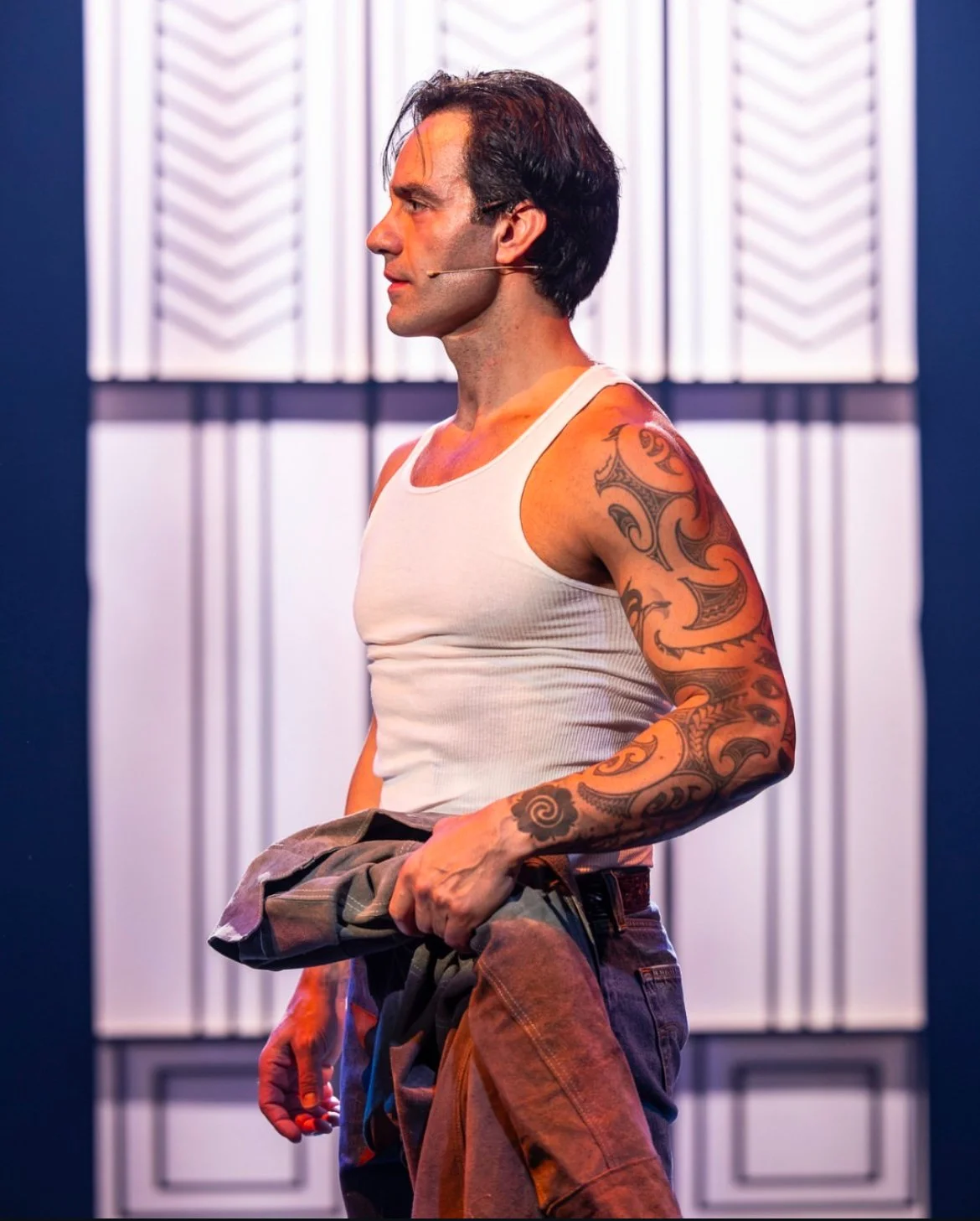 A man with dark hair, tattoos on his right arm, wearing a white tank top, standing in profile on a stage with a blue and white patterned backdrop.