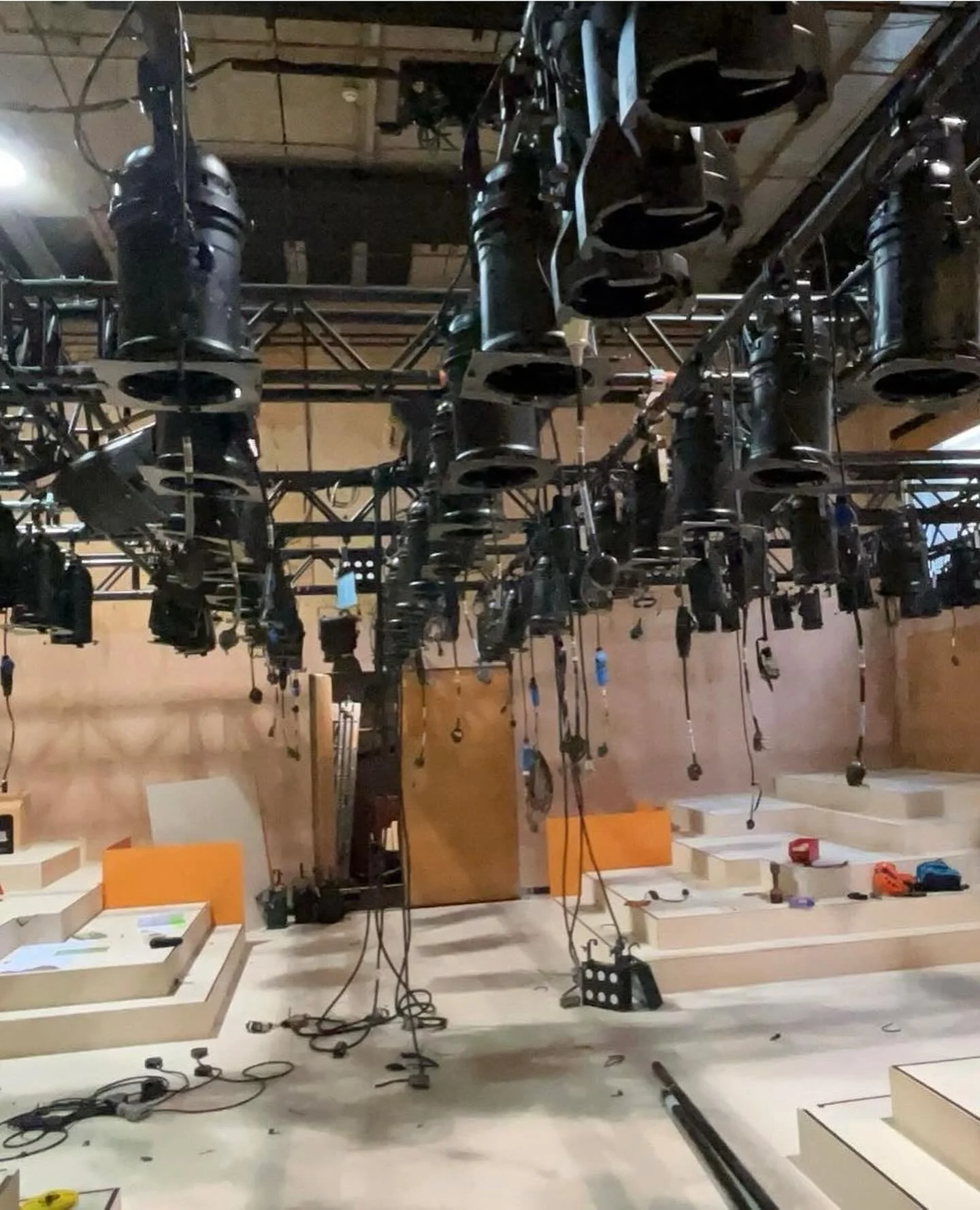 Stage lighting and equipment hanging from the ceiling of a theater or studio. There are stairs and various tools or cables on the floor.