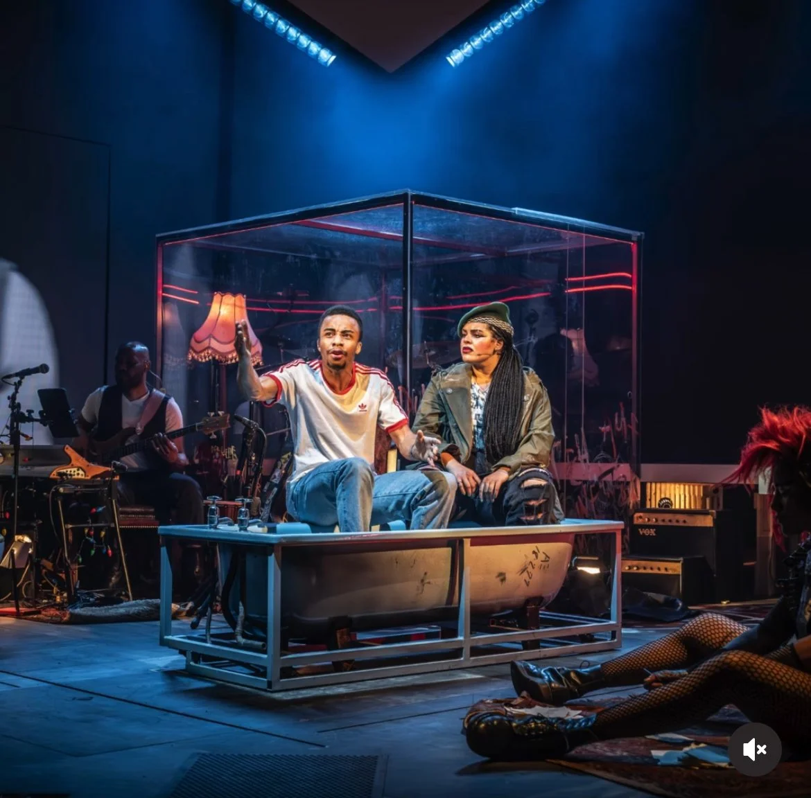 Theatrical performance with two actors seated on a bathtub on stage, surrounded by musicians, in a dark room with colorful neon lighting.