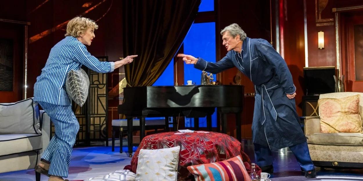 Two actors, a woman in blue striped pajamas and a man in a blue bathrobe, are pointing at each other in a theatrical stage setting. The scene is indoors with a piano, sofas, a stack of cushions, and dramatic lighting.