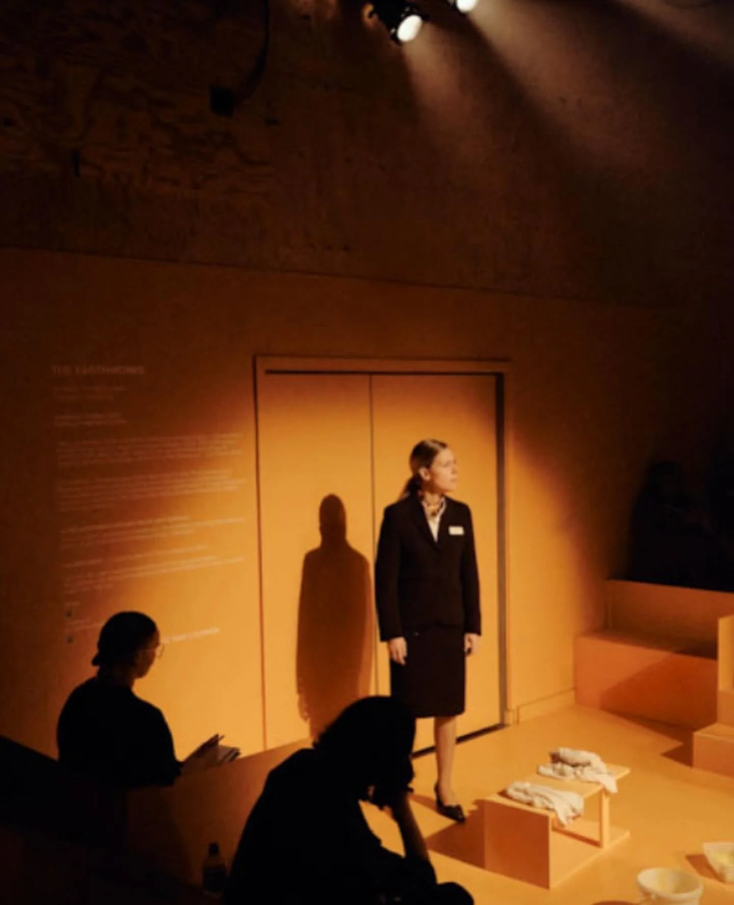 A woman stands on a stage with orange walls, lit by a spotlight. She wears a black blazer, skirt, and a name badge, with a scarf around her neck. In front of her are two seated women, one reading a book, and a table with white cloths and a bowl.