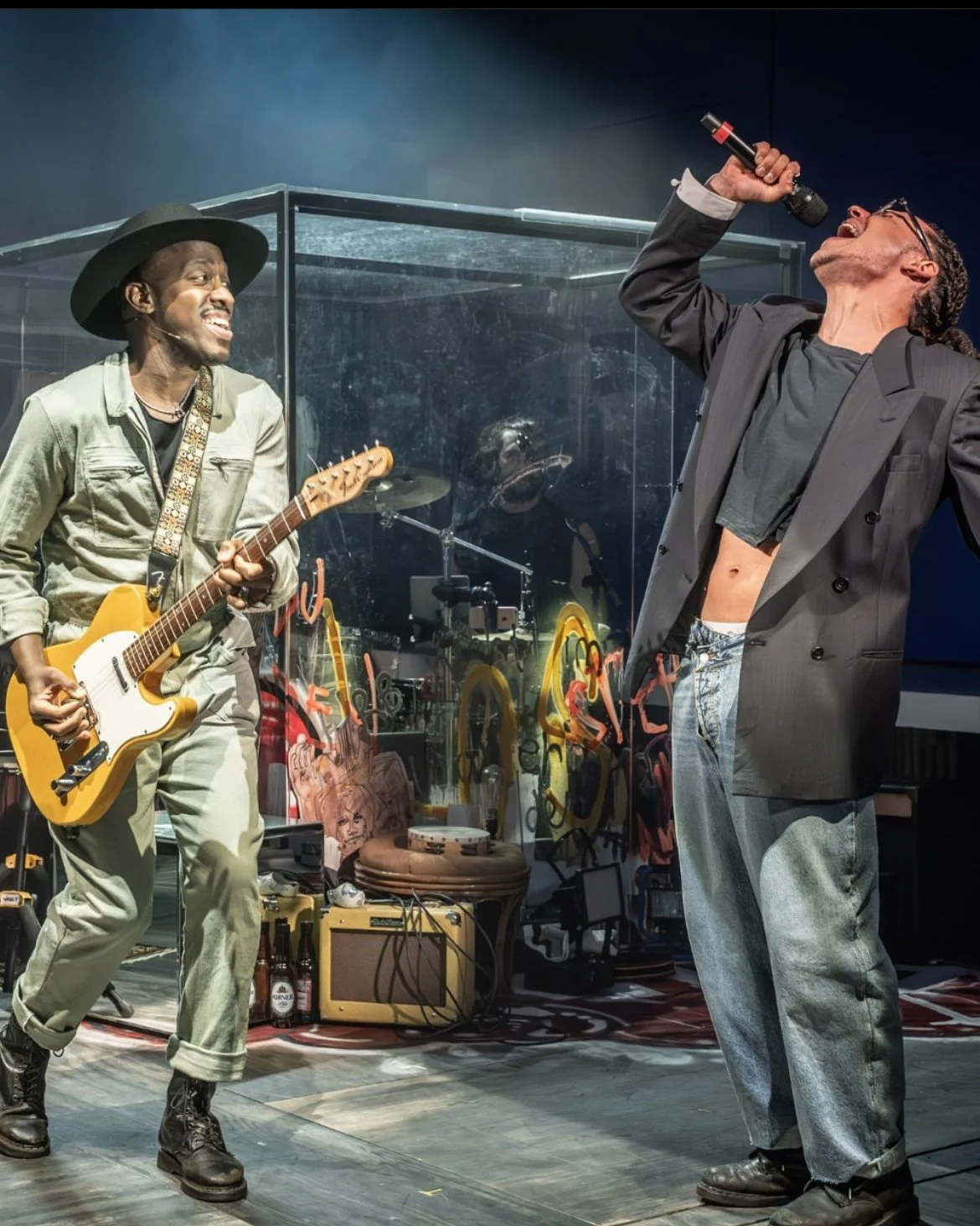 Two musicians performing on stage, one playing guitar and another singing with a microphone, surrounded by musical equipment and graffiti art.