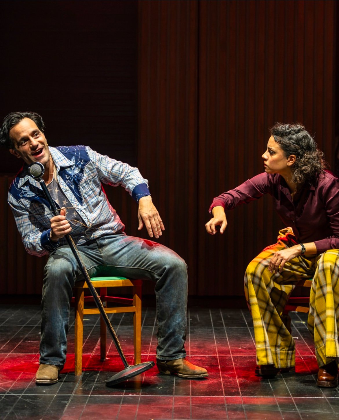 Two actors performing on stage; a man on the left is holding a microphone stand and speaking animatedly, wearing a plaid shirt and jeans, while a woman on the right is listening intently, leaning forward, dressed in a maroon shirt and yellow plaid pa