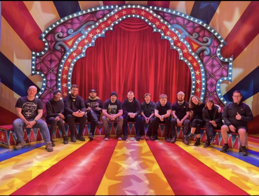 Group of twelve people sitting on benches in front of a large red curtain inside a colorful circus-style theater with bright lights, stars, and striped patterns.