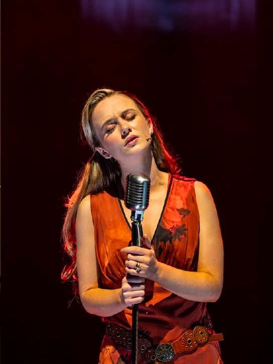 A female singer performing on stage with her eyes closed, holding a vintage-style microphone with both hands, wearing a sleeveless orange dress and a patterned belt.