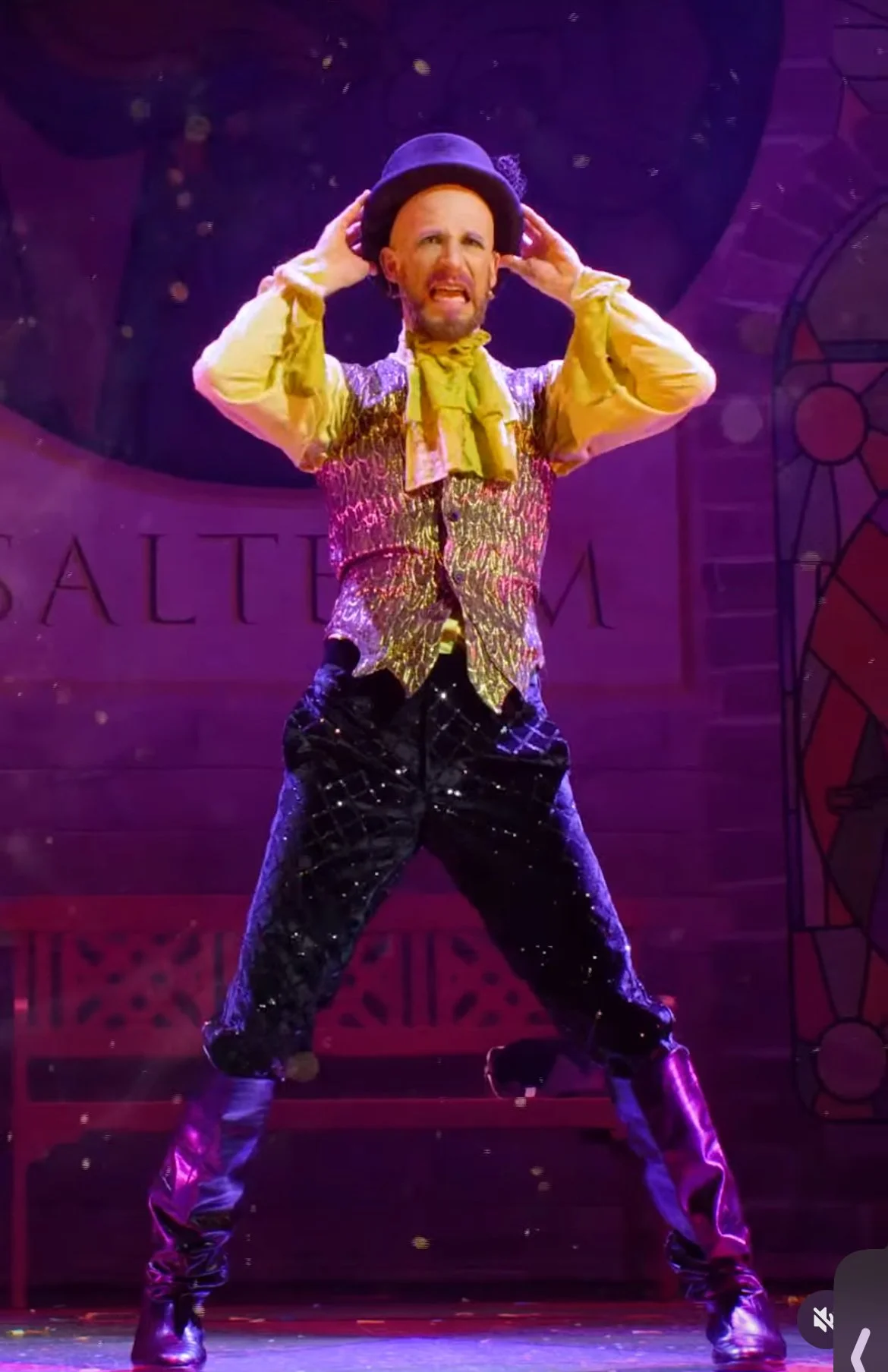 A man performing on stage dressed in a colorful, shiny vest, yellow shirt with a yellow scarf, black shiny pants, purple boots, and a purple hat, with a theatrical expression and stance.