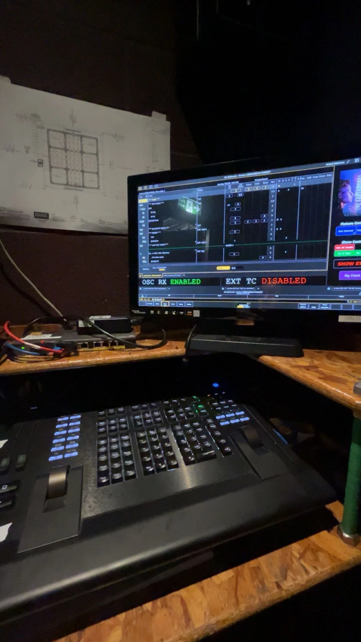 Photo of a computer setup with a monitor displaying a digital audio workstation software, and a control surface with numerous buttons and sliders on a wooden desk in a dimly lit room.