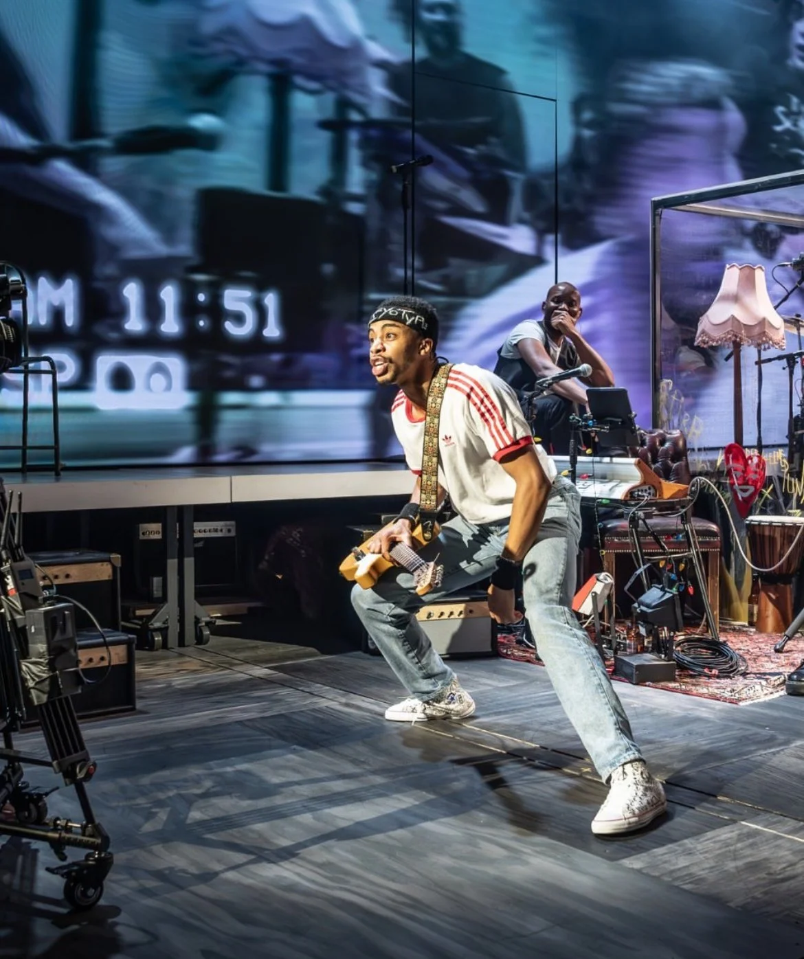 Musician playing guitar and dancing on stage with background video screen and various stage props.