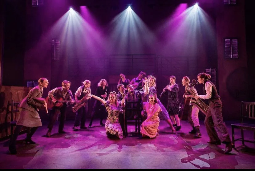A theatrical stage scene with multiple actors, some holding musical instruments, under colorful purple and pink stage lighting, creating a lively and cheerful atmosphere.