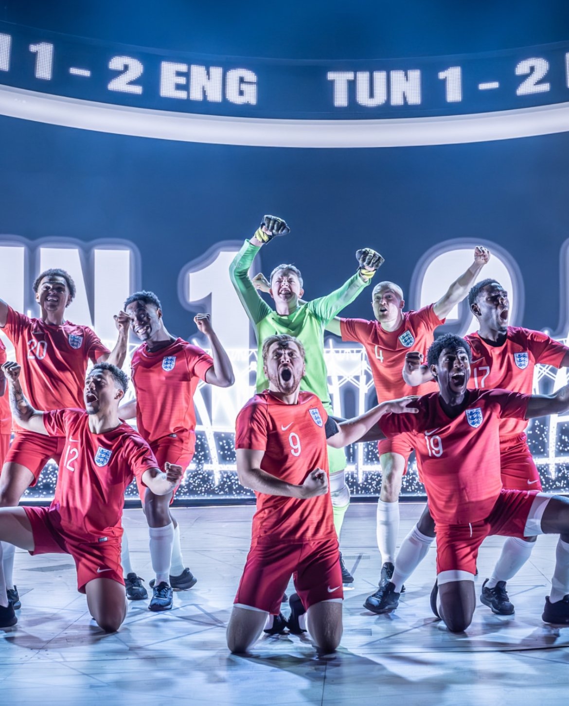 A group of seven male soccer players in red jerseys and black shorts celebrating on stage. They are kneeling and standing with their fists raised, with a large digital scoreboard or display in the background showing the score and text.