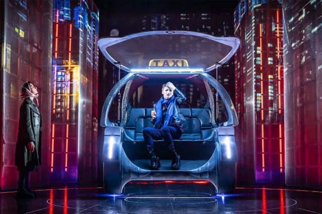 A futuristic taxi stand with a woman sitting inside a transparent, bubble-like taxi surrounded by neon lights, while another woman stands outside at night.