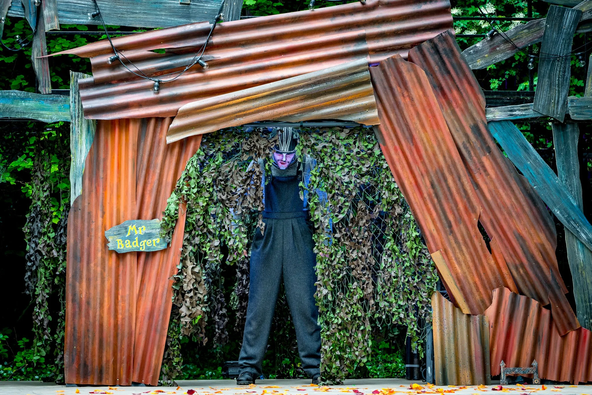 A theatrical stage with an actor dressed as a badger, standing behind a rustic, handmade set with a sign labeled 'Mr Badger', surrounded by green foliage and autumn leaves on the ground.