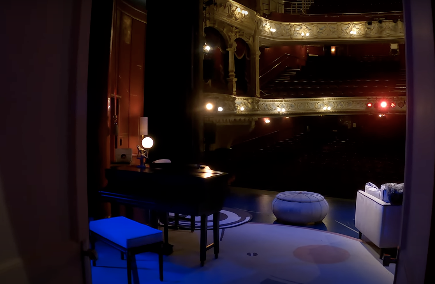 Interior view of a theater stage with elaborate gold and white balustrades, a small living room setup with a desk, lamp, piano, and seating, seen through a window or open door.