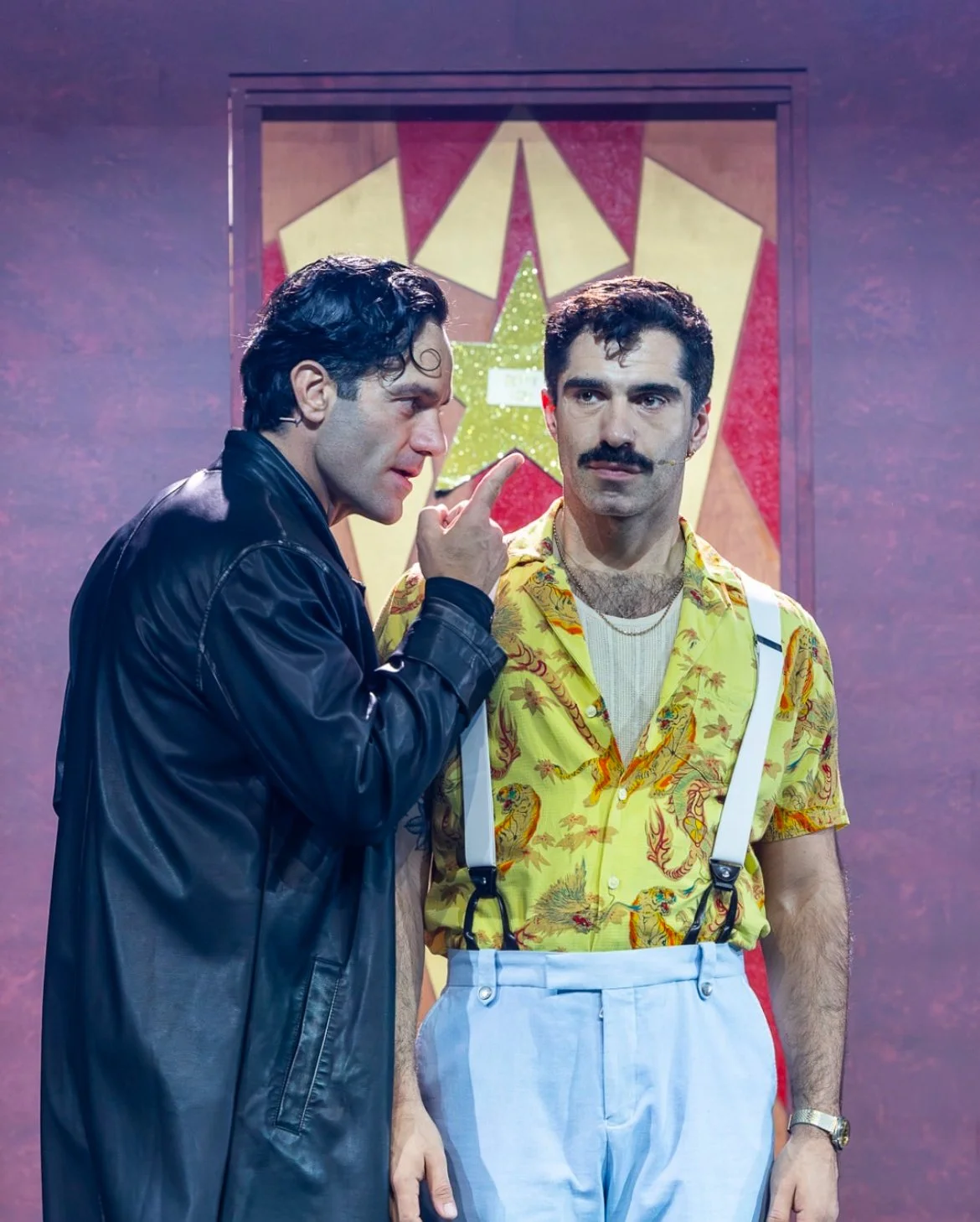 Two men standing in front of a colorful star-themed backdrop, one man wearing a leather jacket talking and pointing his finger near the other man's face, who is wearing a yellow tropical shirt with suspenders.