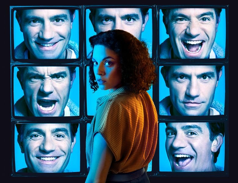 A woman with curly hair and a checkered shirt standing in front of a display of multiple television screens showing various expressive faces.