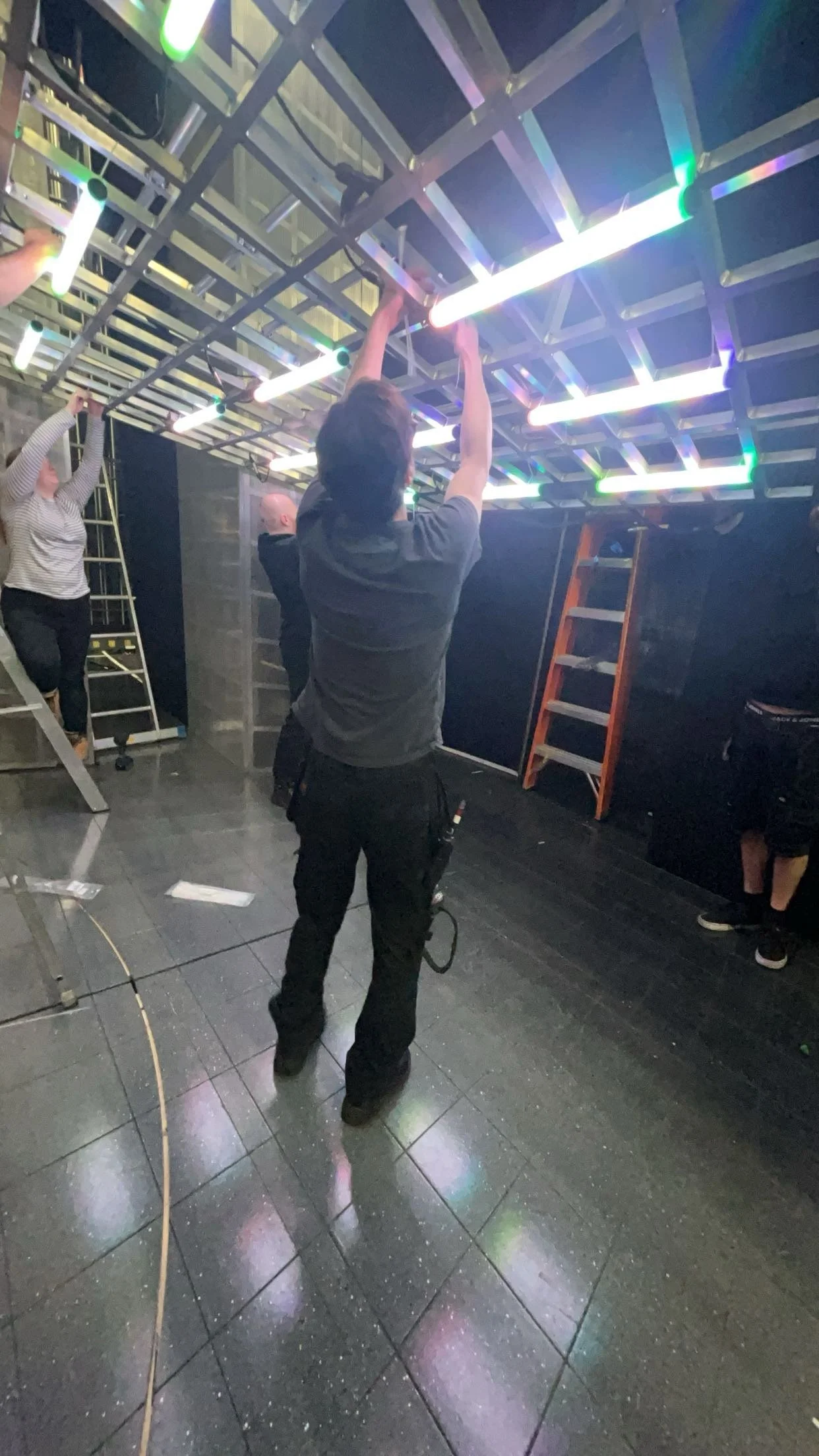 People installing colorful LED light fixtures on a ceiling structure with ladders in a dark room.