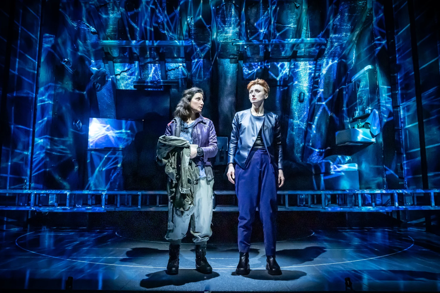 Two women in futuristic clothing stand on a stage with a high-tech, blue-lit set behind them, possibly in a science fiction theater production.