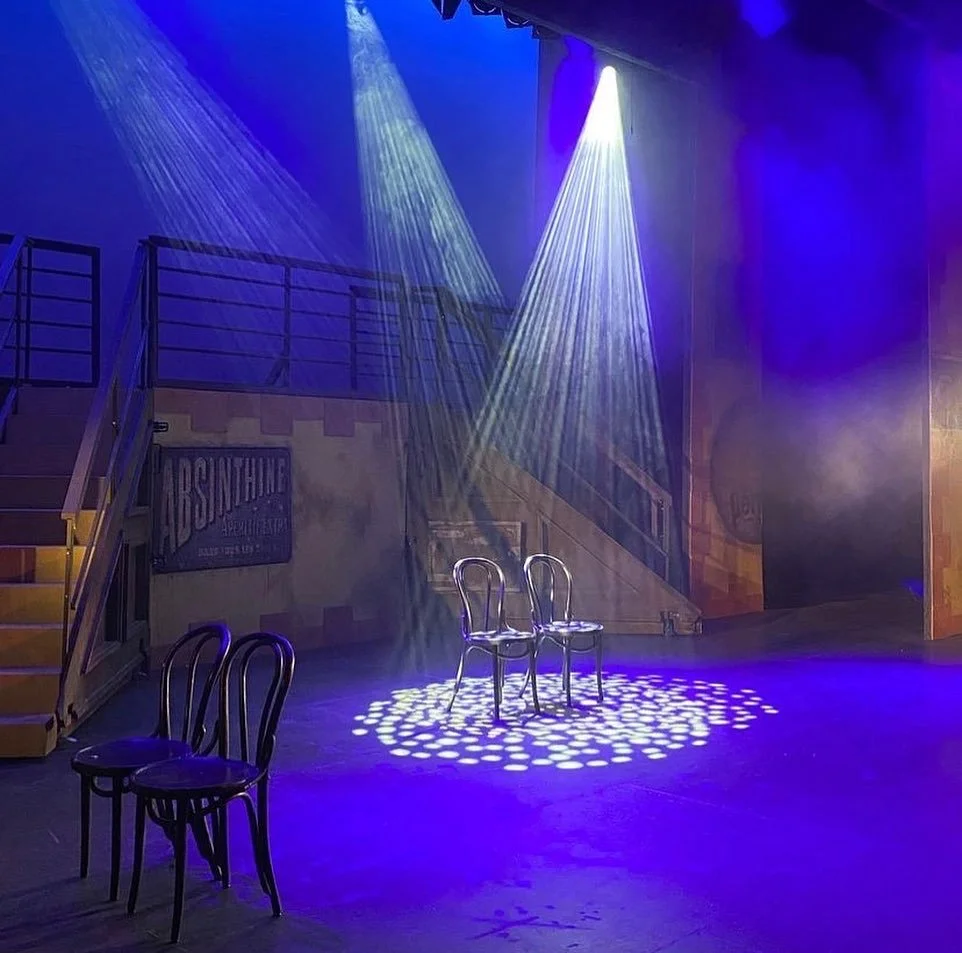 The image shows a stage with two chairs under bright spotlights, creating illuminated circles on the floor. Two additional chairs are positioned in the foreground, while a staircase and a sign reading "ABSINTHE" are visible in the background. The sce
