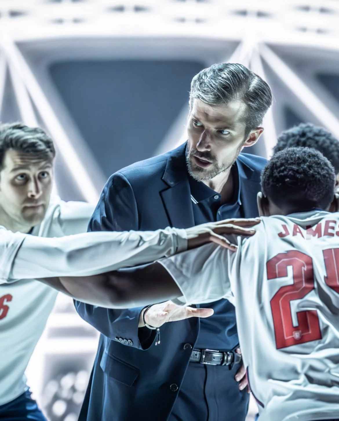 A coach in a dark suit passionately shouting at his soccer team during a game, with players in white jerseys around him.