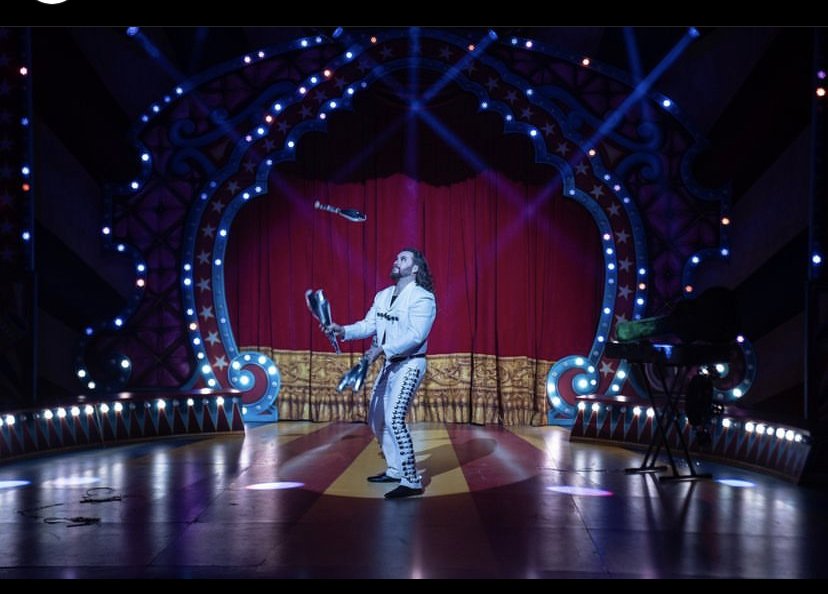 A magician in a silver suit performs on a colorful stage with red curtains, holding juggling clubs, while a microphone is suspended in the air and a keyboard is on the right side of the stage.