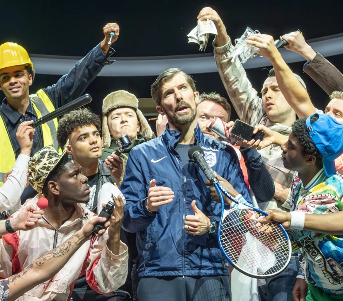 A man in a blue jacket, holding a microphone, stands at the center surrounded by diverse individuals, some holding microphones, smartphones, and tennis racquets, in a press conference setting.