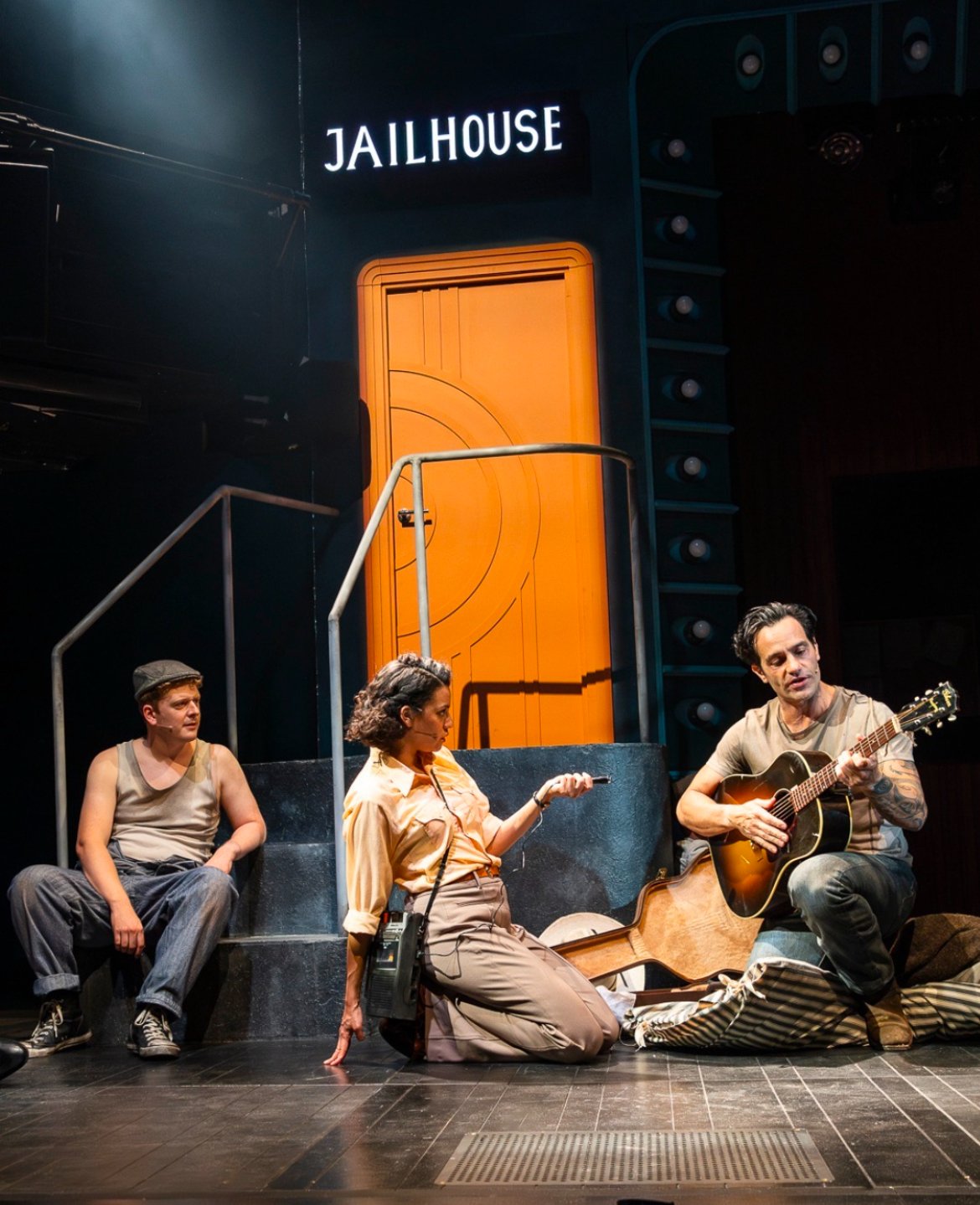 A theatrical stage scene labeled 'Jailhouse' with four actors. One actor on the left is sitting, the middle actor is kneeling, and the actor on the right is playing guitar. The set includes a large orange door and a sign that reads 'Jailhouse'.