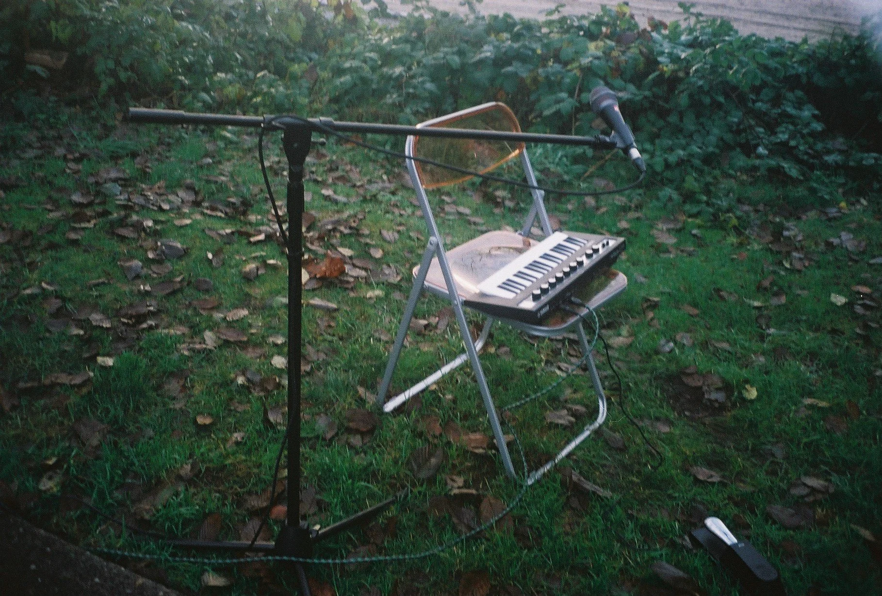 A microphone stand and a small musical keyboard on a metal chair outdoors on a grassy area with fallen leaves.