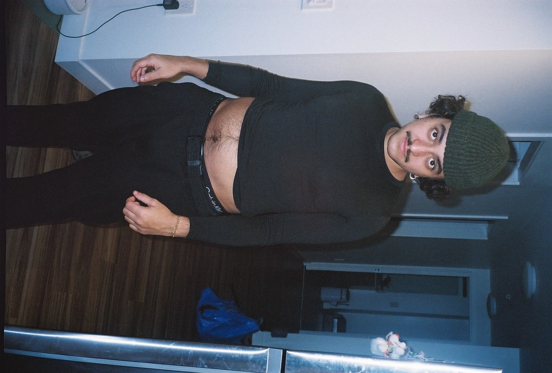 A woman with short curly hair, wearing a green beanie, black long-sleeve top, and black pants, standing in a room with dark wood flooring, a white wall, and a white door in the background.