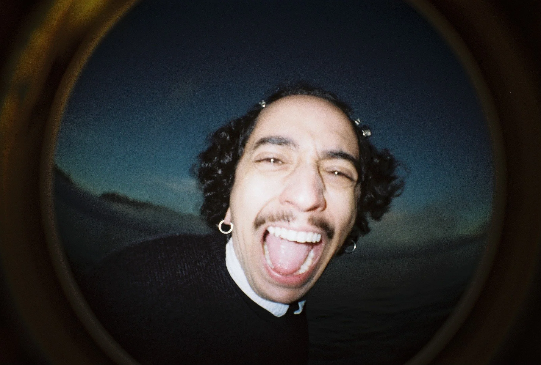 Close-up of a man with dark, curly hair and earrings, smiling with his mouth open, taken with a fisheye lens against a twilight sky.