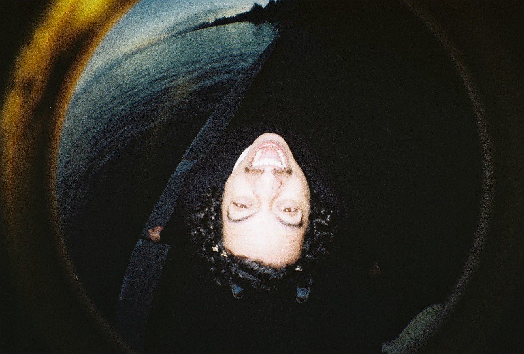 A woman with curly hair and glasses sticking out of her pocket, smiling with her mouth open, looking into a fisheye lens, with a body of water and a sunset in the background.