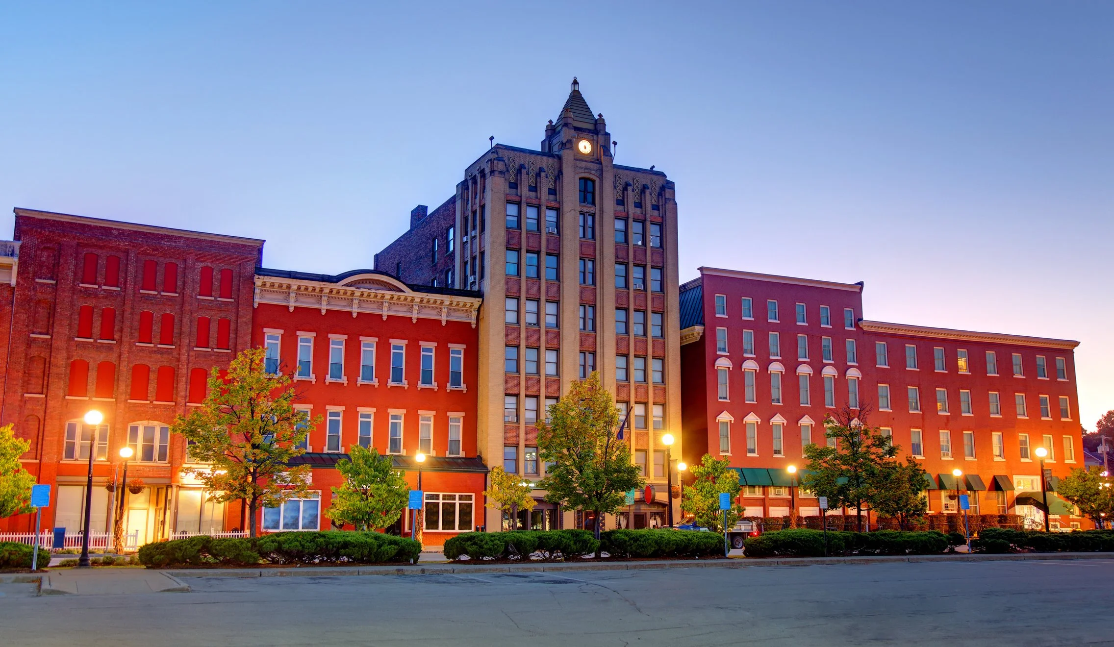A city street scene at dusk with a row of red brick buildings and a central taller building with a clock tower, street lamps, trees, and parked cars.