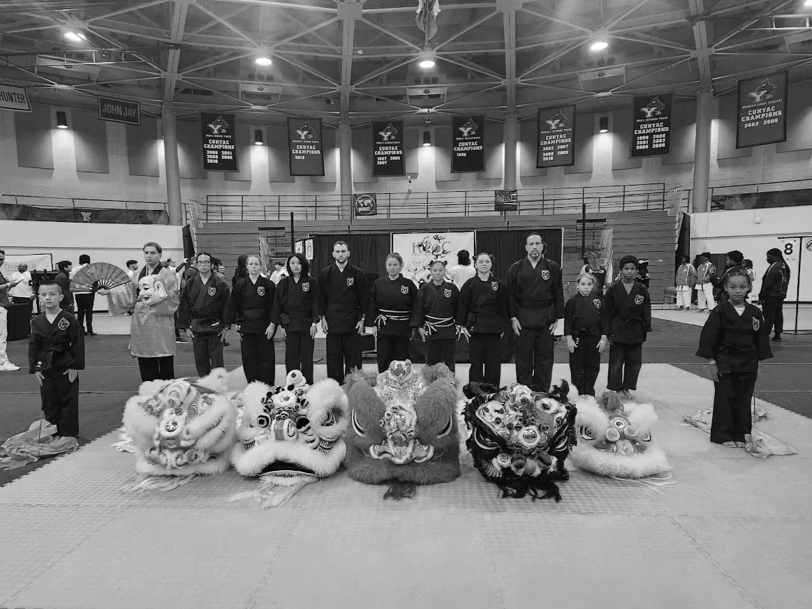 Group of martial artists standing in a gymnasium with lion dance costumes in front of them, banners hanging from the ceiling celebrating championships, and people in the background.