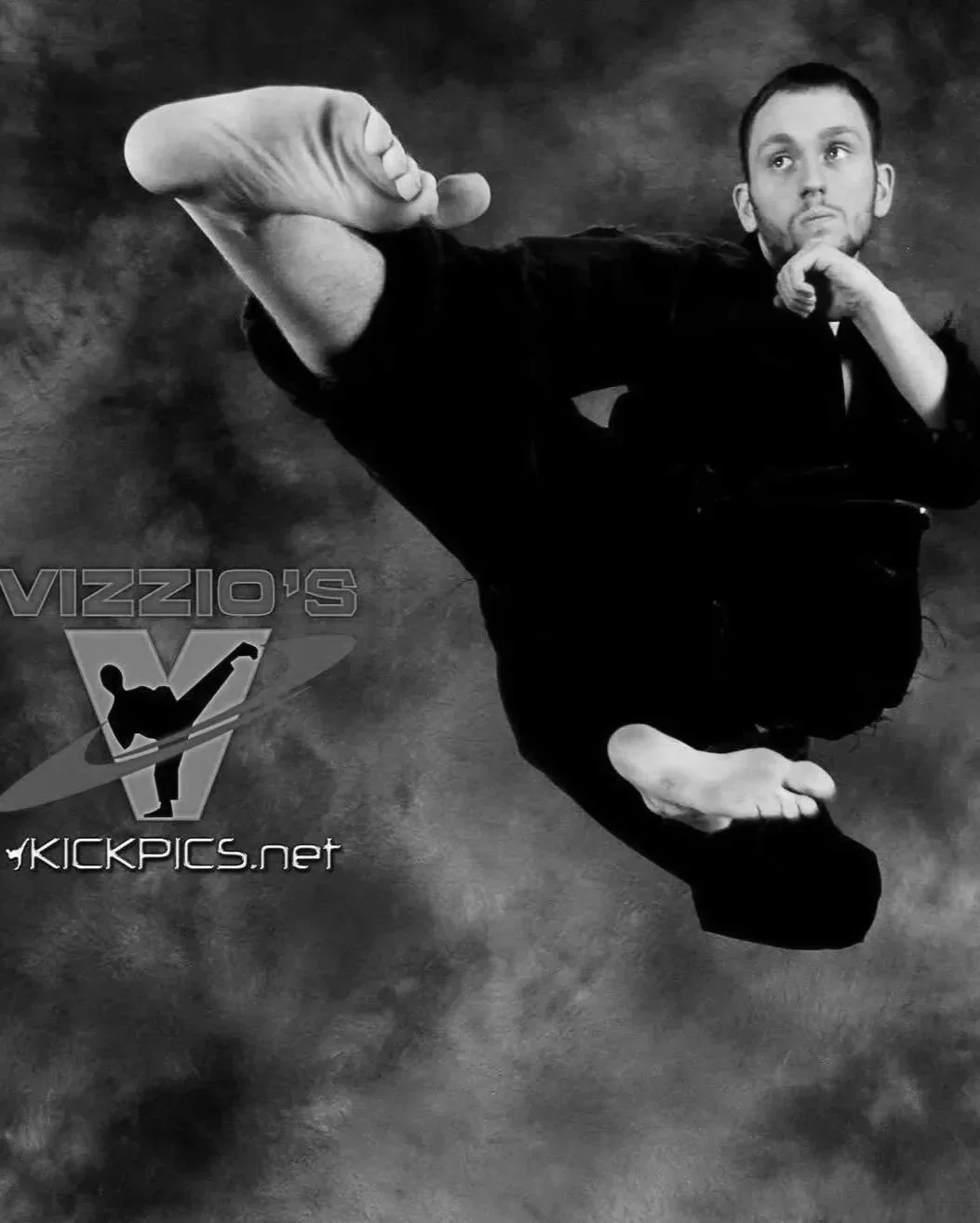 A man in black clothing is performing a flying kick in a martial arts pose, with his right leg extended and his left arm supporting his balance. The background is a dark, cloudy texture, and there is a logo in the lower left corner that says 'Vizzo's