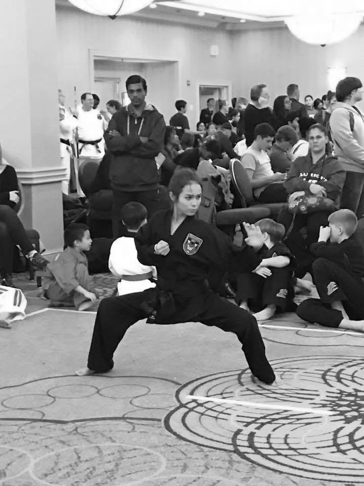 A young martial artist demonstrates a stance in front of a crowd at a martial arts event or competition, with many children and adults watching in the background.