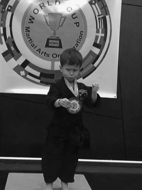 Young boy in a martial arts uniform holding a medal in front of a large banner with a trophy and the text 'World Cup'.
